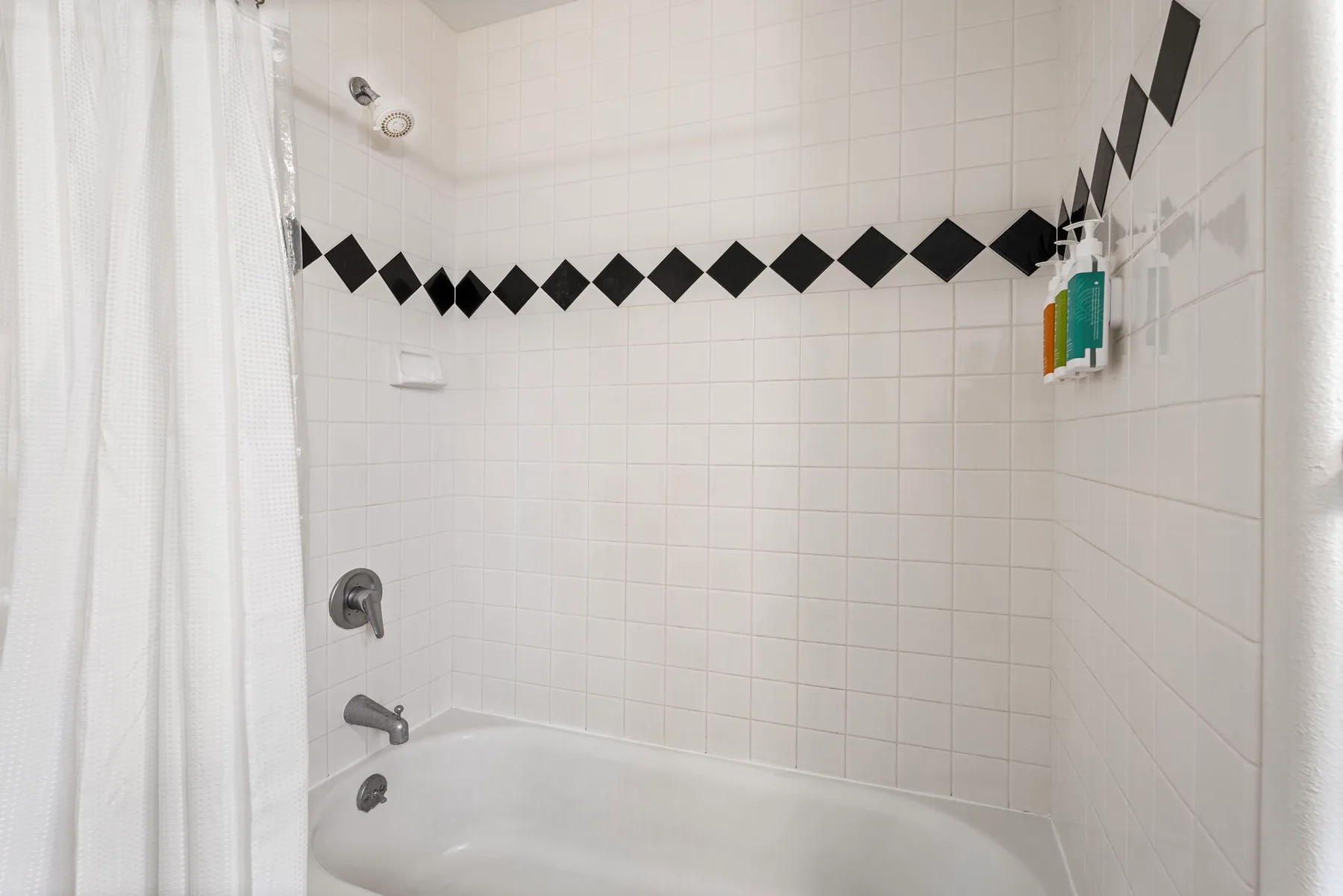 Baño alicatado con ducha y bañera, cortina blanca y detalles de rombos negros.