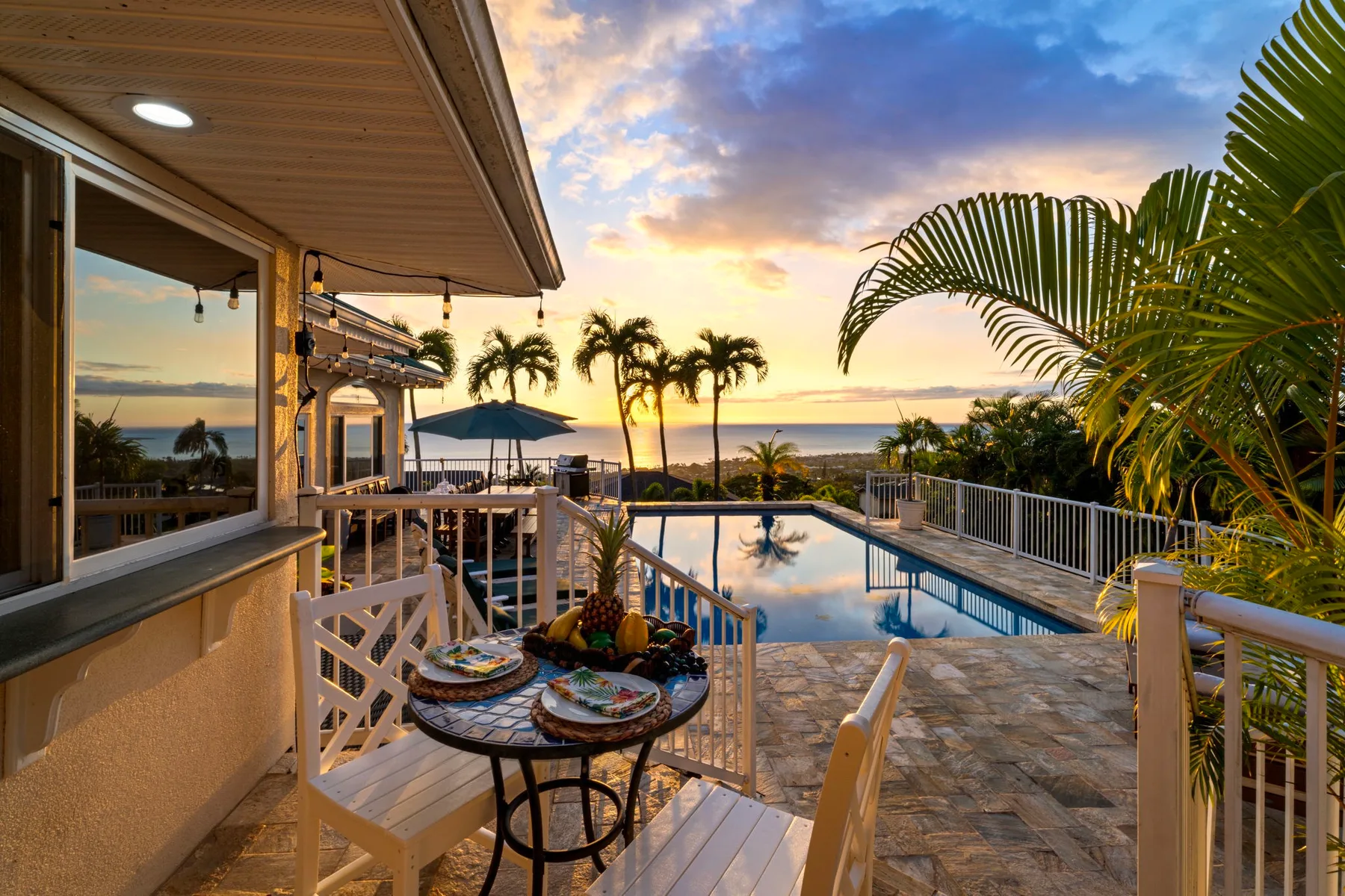Un patio cubierto da a una piscina, palmeras y el océano al atardecer.