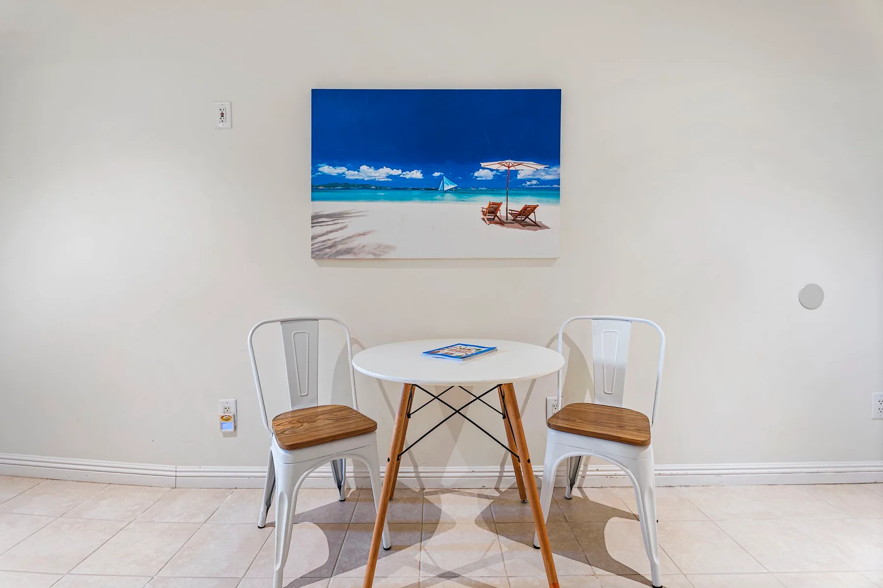 Un pequeño comedor con una mesa redonda y dos sillas bajo un cuadro de playa.