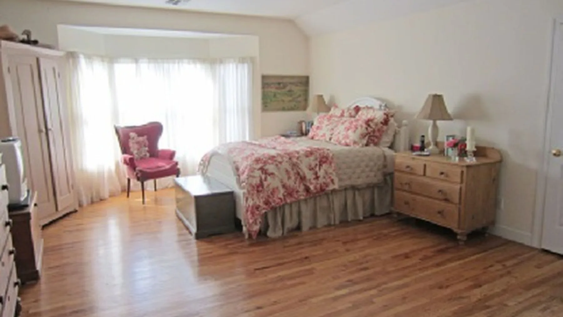 A bedroom with a floral-covered bed, a wooden dresser, and a red armchair.