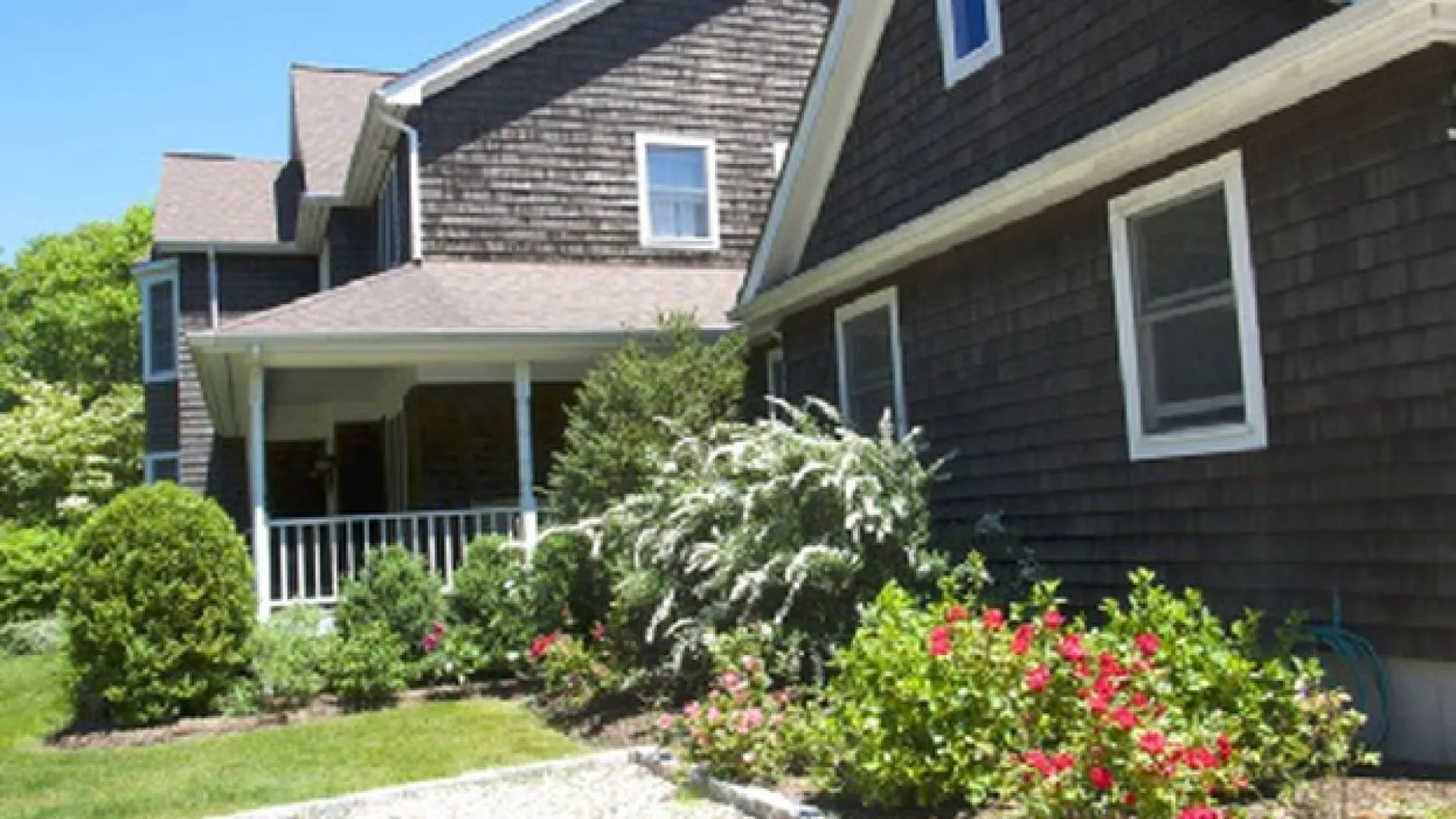 A dark-shingled house exterior with a covered porch and flowering garden.