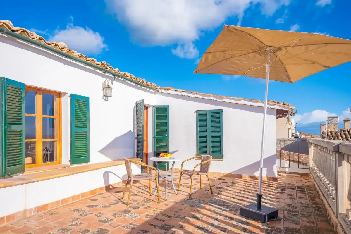 A sunny rooftop terrace with a white building, green shutters, and outdoor seating.