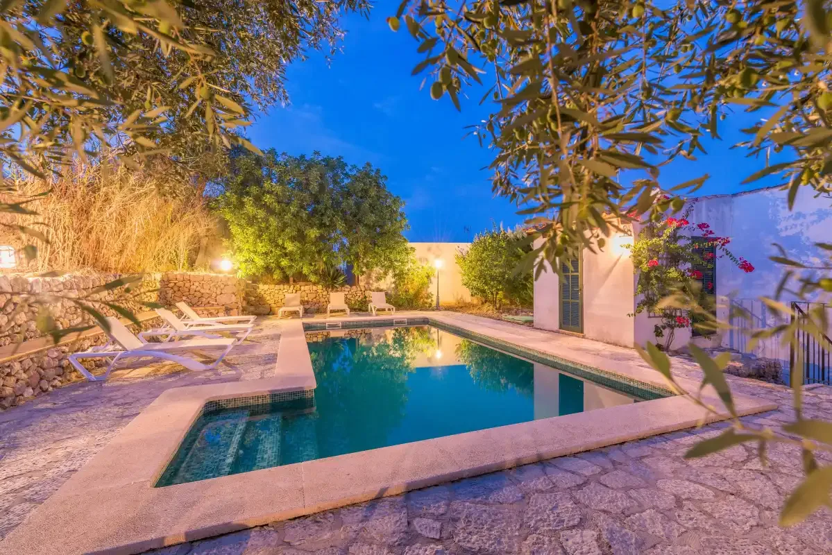 An outdoor swimming pool sits among stone paving, lounge seating, and leafy trees.