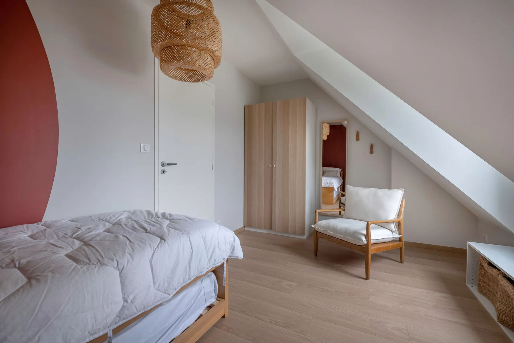 A bedroom with a bed, wooden wardrobe, and cushioned chair beneath a sloped ceiling.