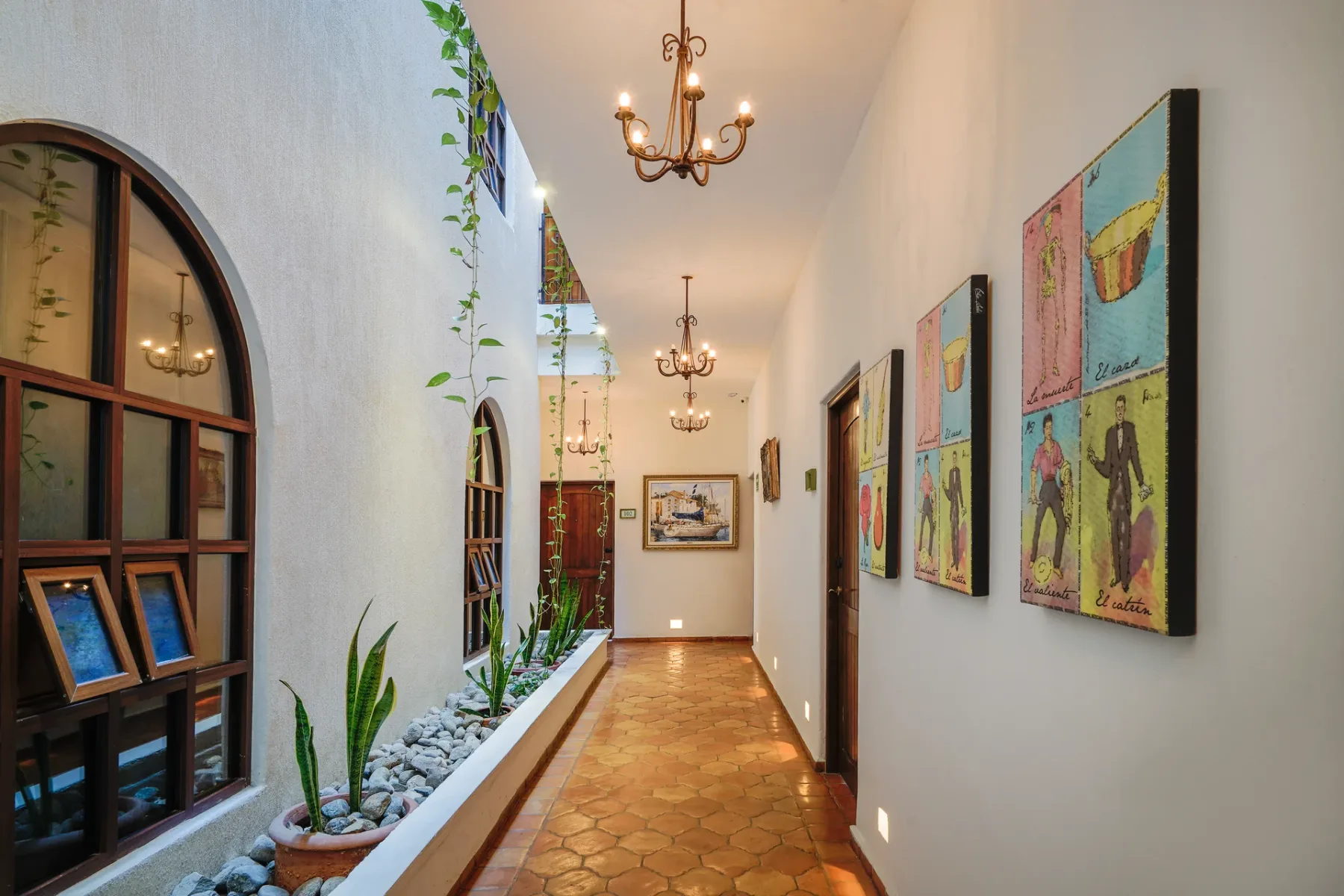 Pasillo del hotel con puertas de madera arqueadas, plantas en macetas y obras de arte coloridas.