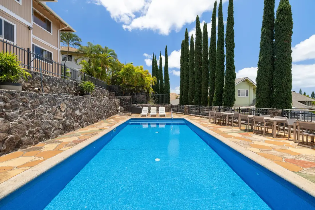 A swimming pool sits beside a patio with outdoor seating and tall trees.