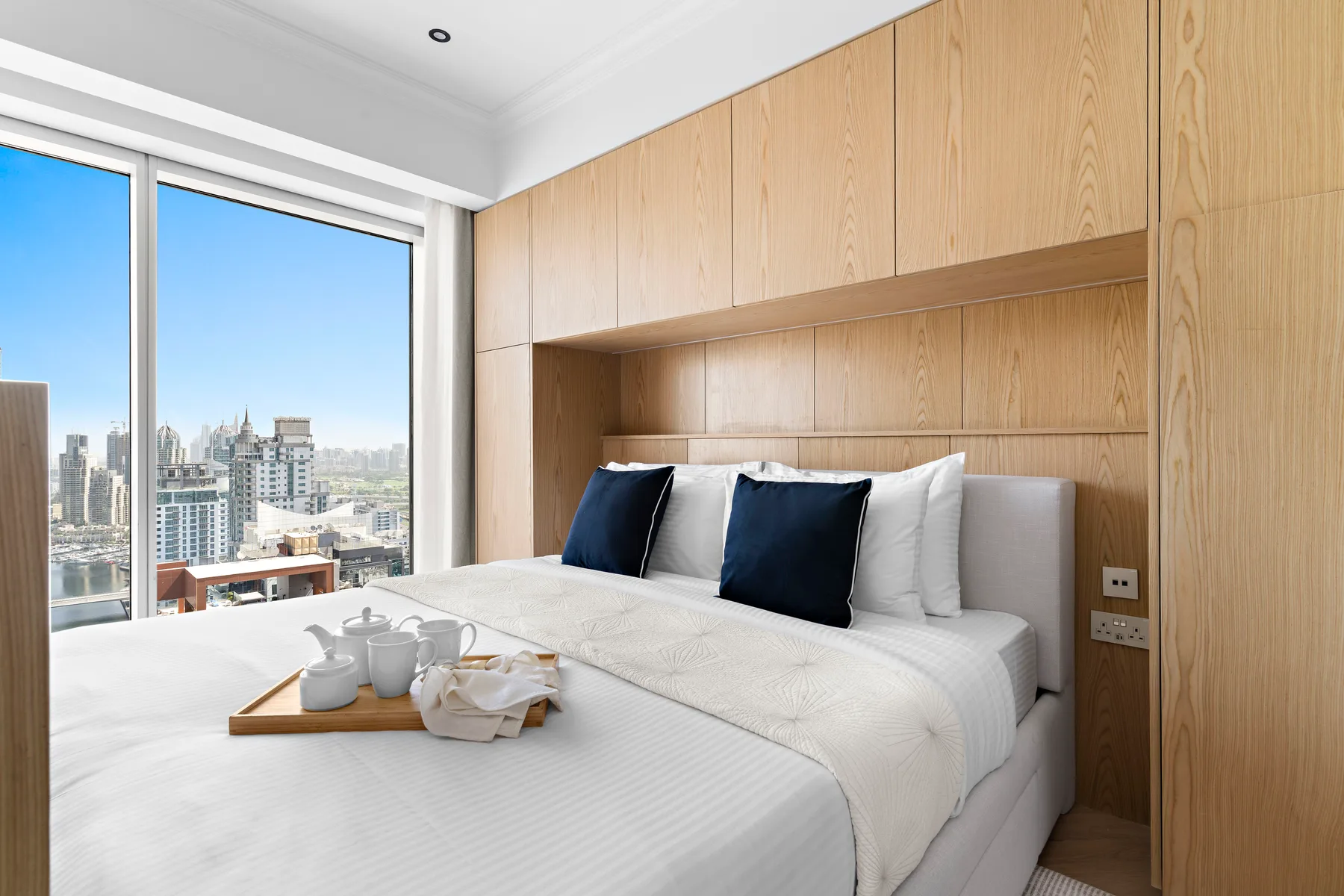 Un dormitorio con una cama bien tendida y grandes ventanas con vistas a edificios de la ciudad.