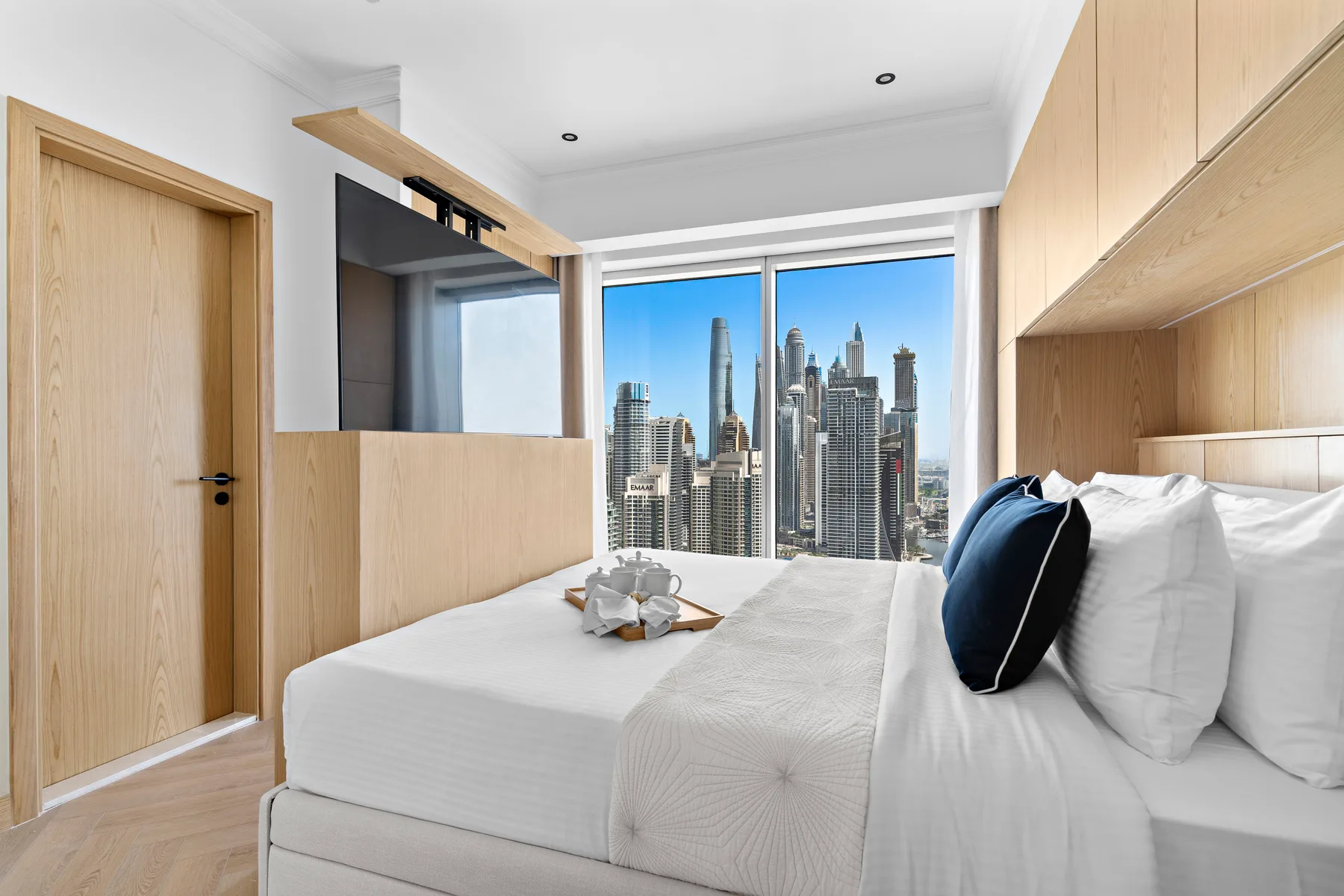 Un dormitorio luminoso con una cama tendida y vistas de la ciudad por grandes ventanas.