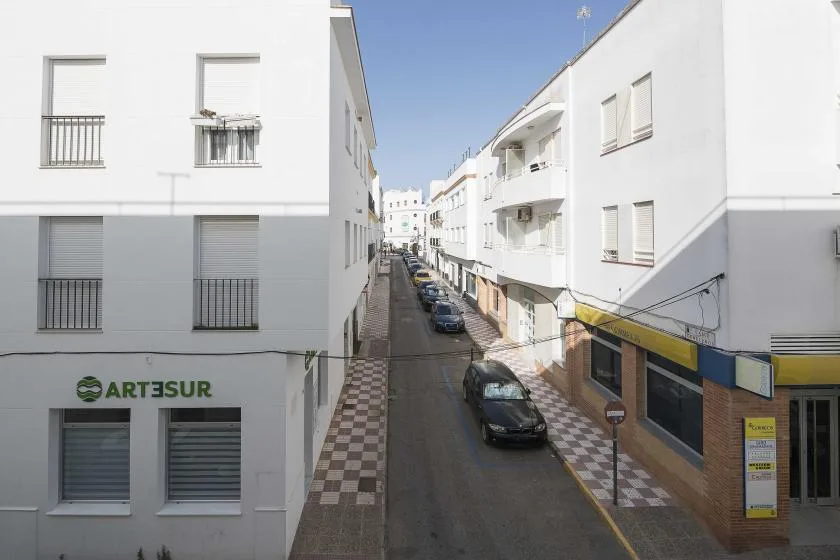 A narrow street runs between white buildings, with parked cars along the road.