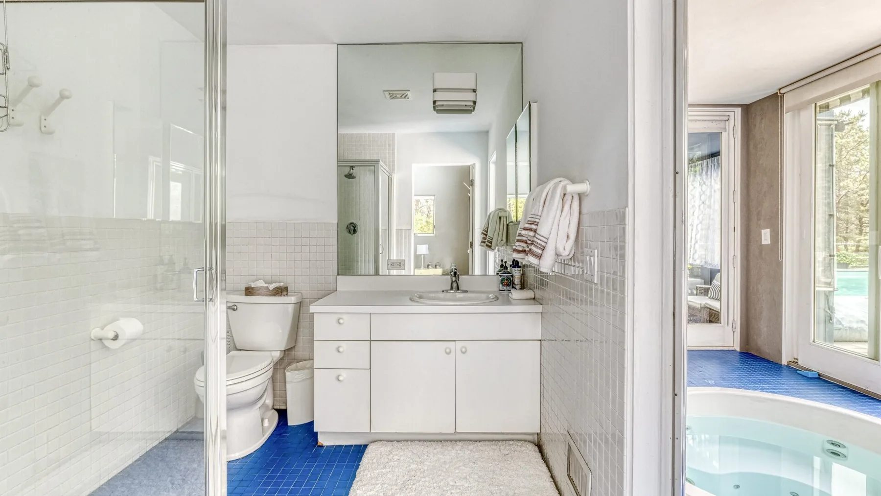 A bathroom with a glass shower, vanity, and mirror opens onto a room with a spa tub.