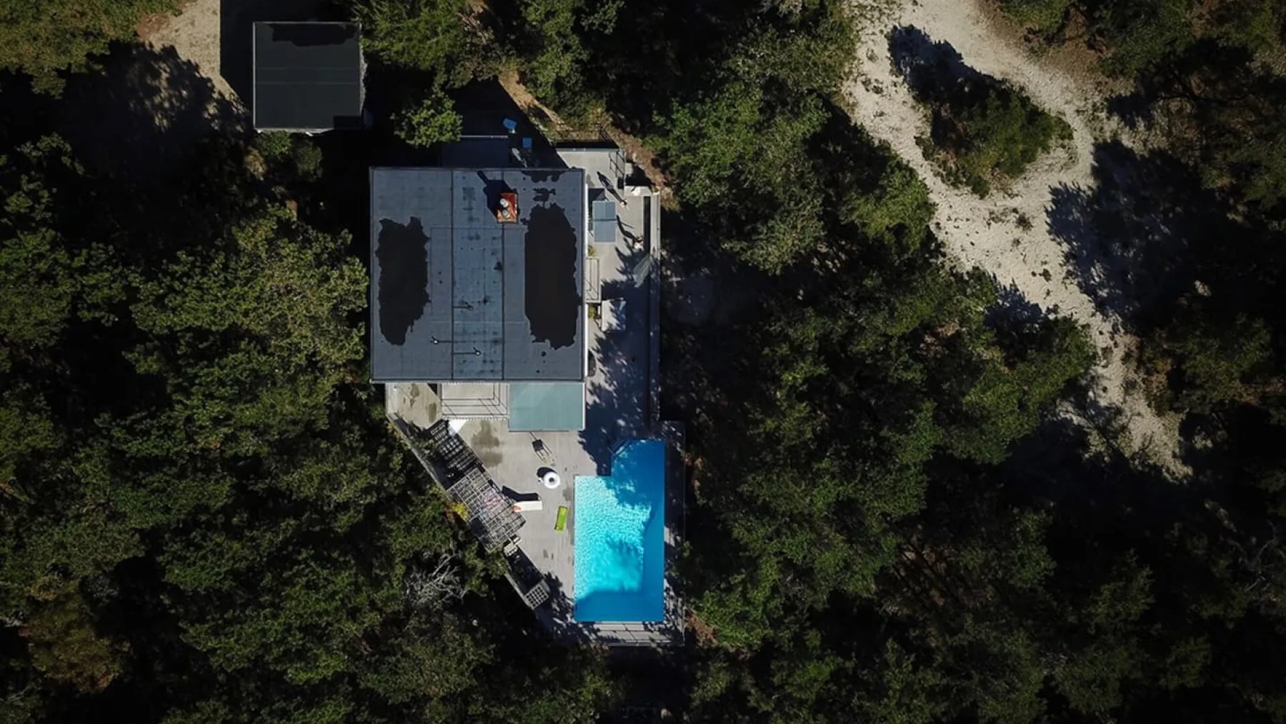 Aerial view of a building beside a bright blue swimming pool, surrounded by trees.