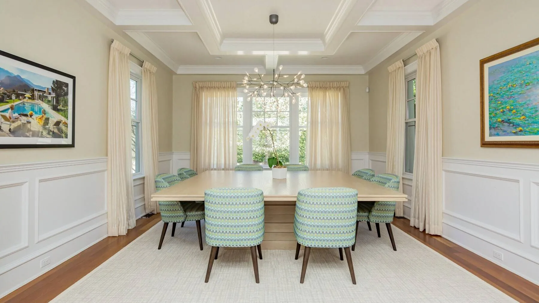 A bright dining room with a long table, upholstered chairs, and large windows.