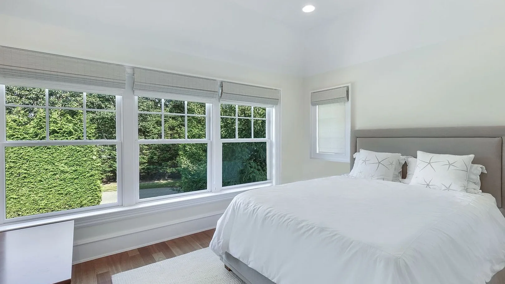 A bright bedroom with a white bed beside wide windows overlooking greenery.