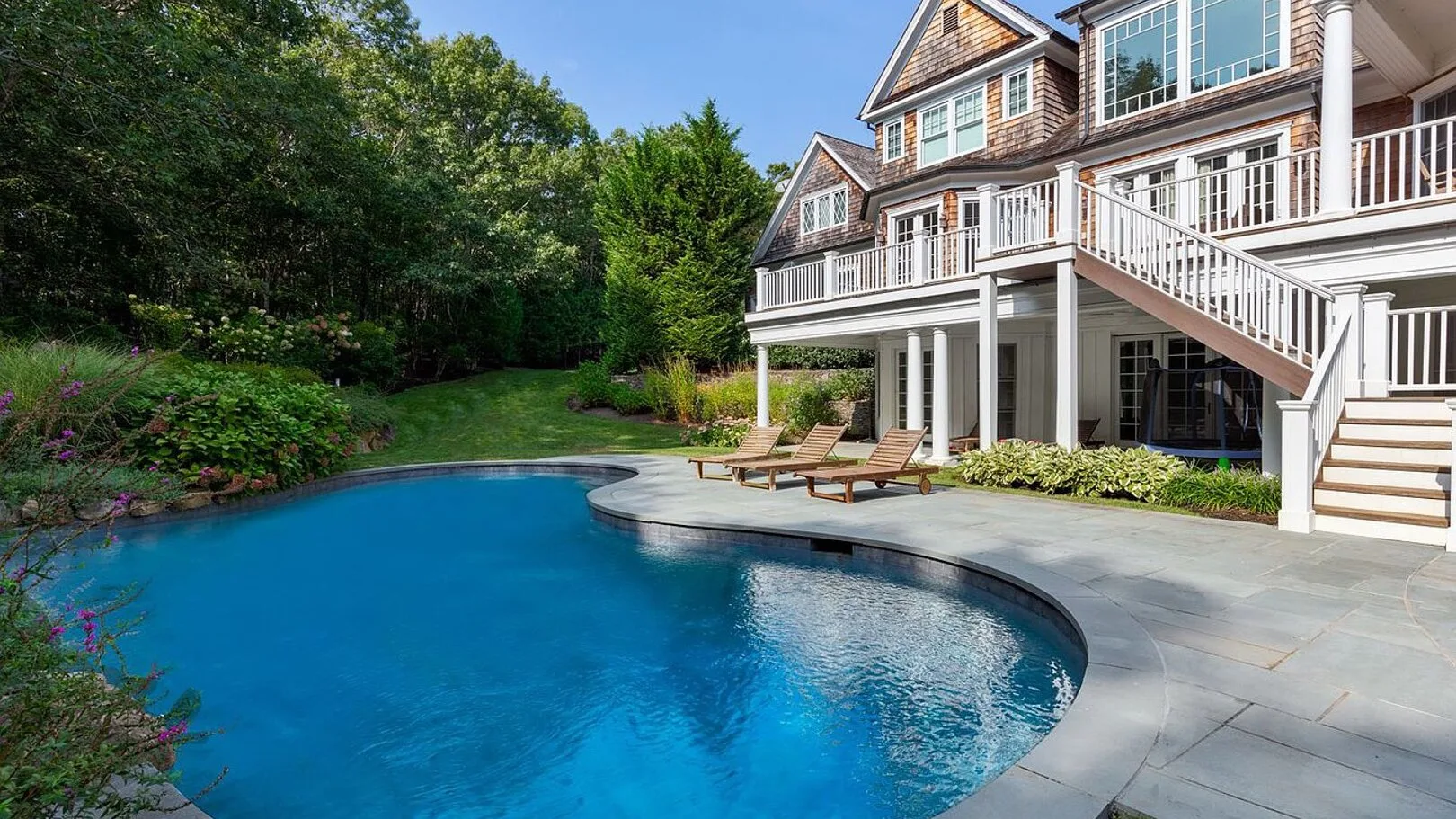 A backyard pool beside a large house, with a patio and surrounding greenery.