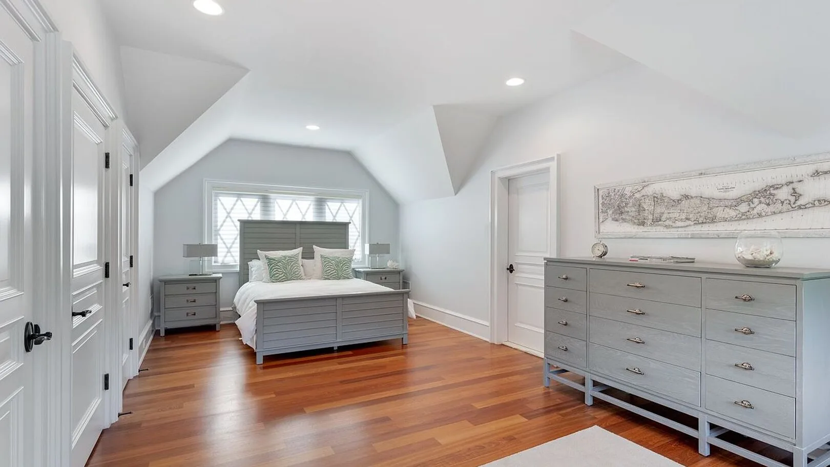 A bright bedroom with a bed, gray dressers, and polished wood flooring.