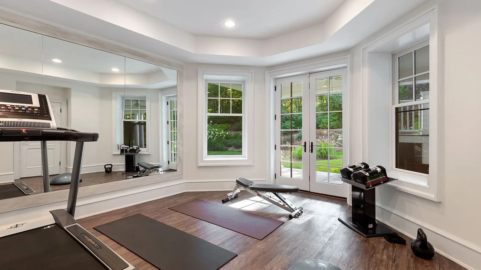 A home gym with a treadmill, exercise mat, and weight bench beside large windows.