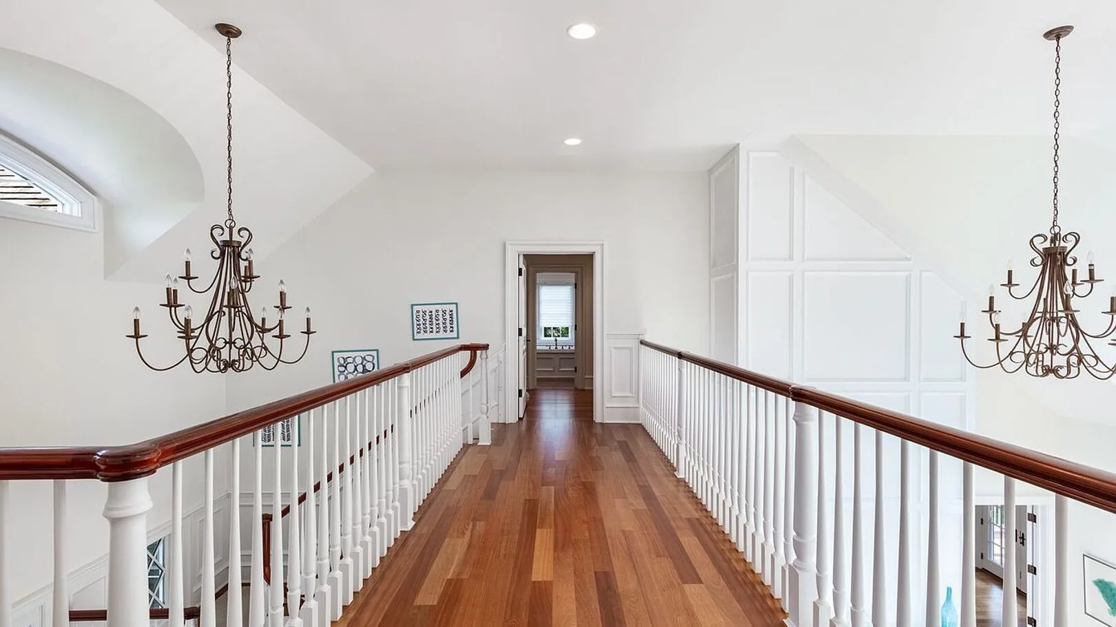 A bright upstairs landing with wood flooring, white railings, and hanging chandeliers.