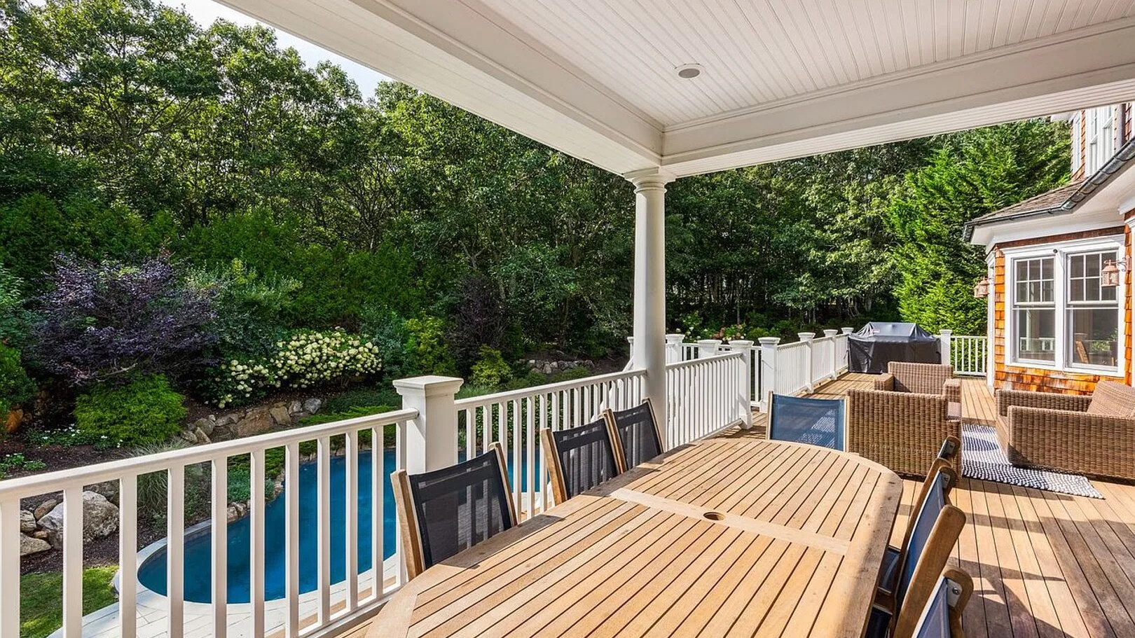A covered wooden deck overlooks a swimming pool and leafy garden.