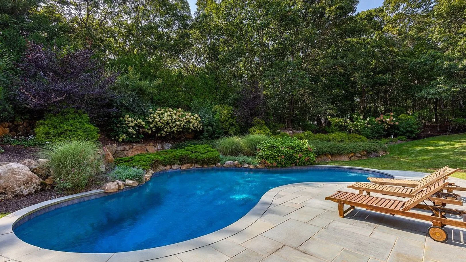 An outdoor swimming pool with paved decking, lounge seating, and surrounding greenery.