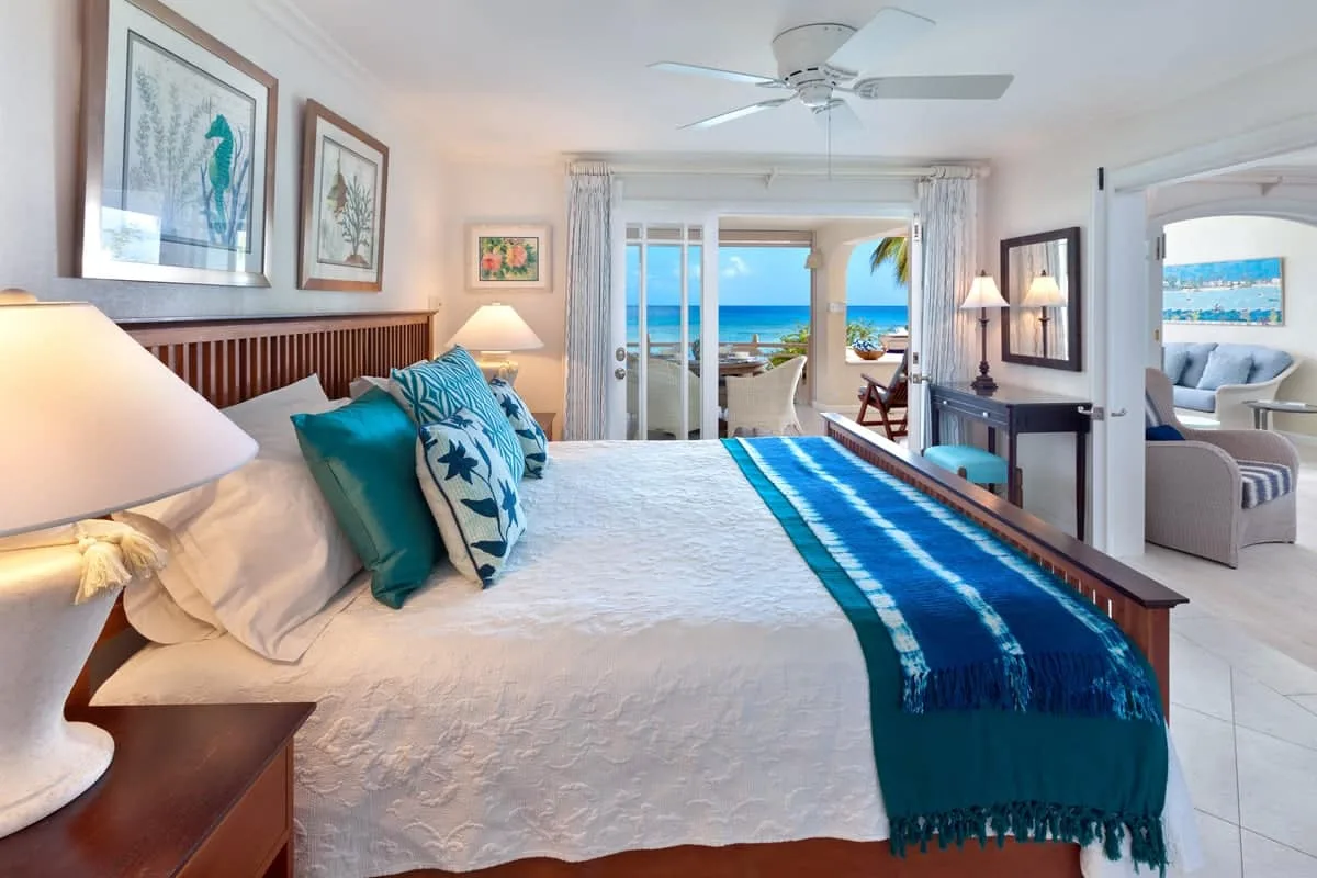 A bright bedroom has a large bed with blue bedding and ocean views through the balcony doors.