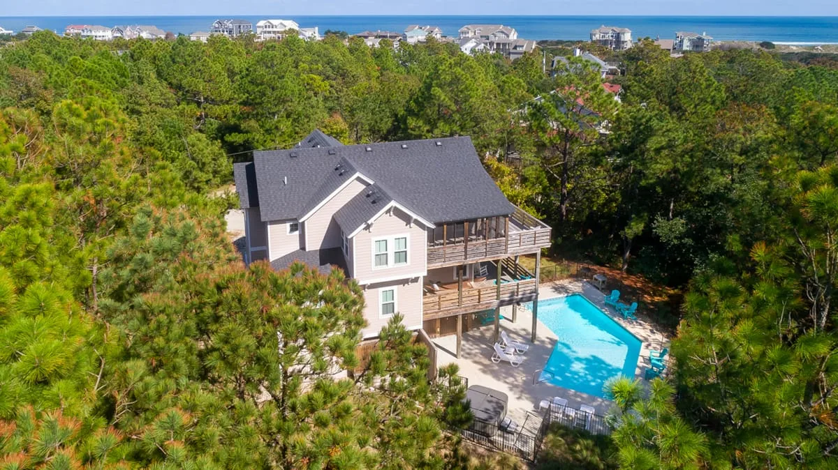 A large house stands among trees beside a bright blue swimming pool.