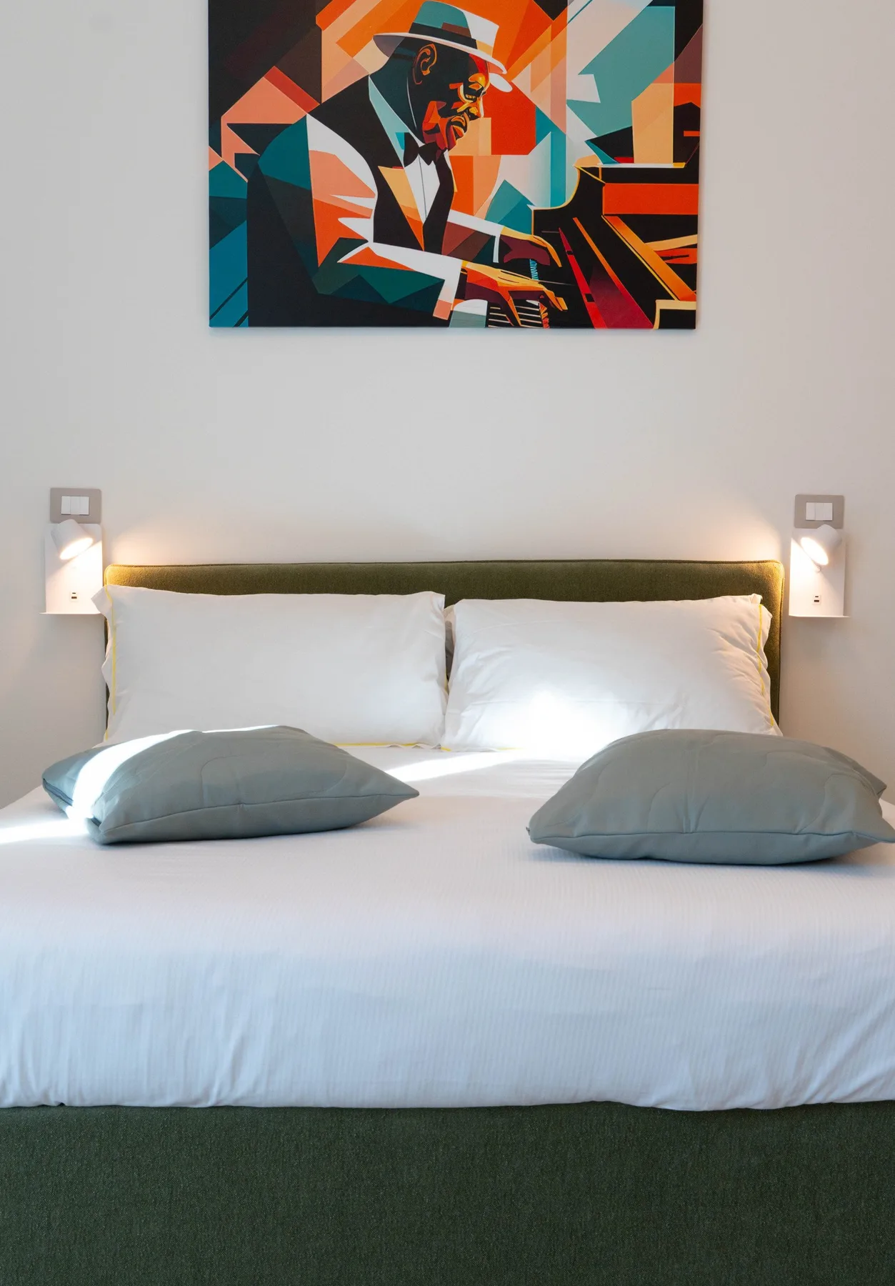 A neatly made bed sits beneath colorful artwork on a light wall.