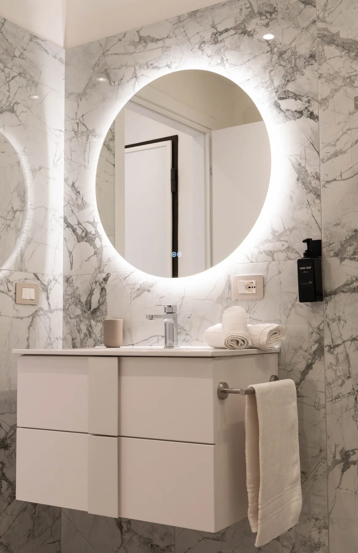 A bathroom vanity with a round illuminated mirror and a towel.