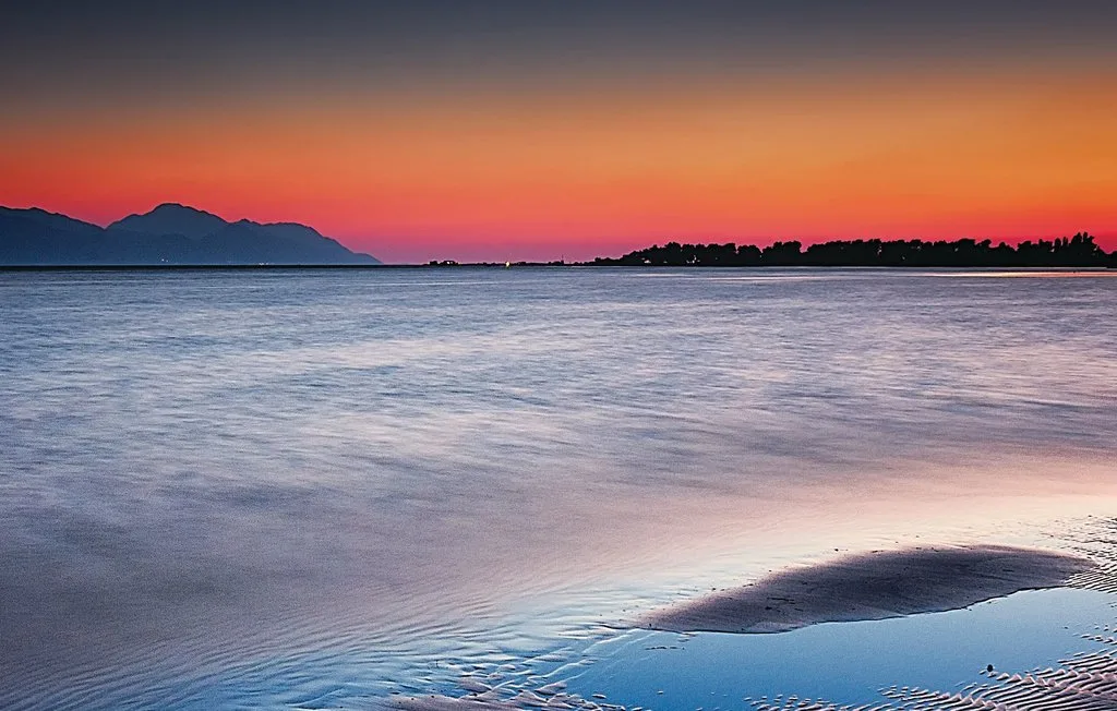 Una orilla tranquila al atardecer, con montañas lejanas bajo un cielo colorido.