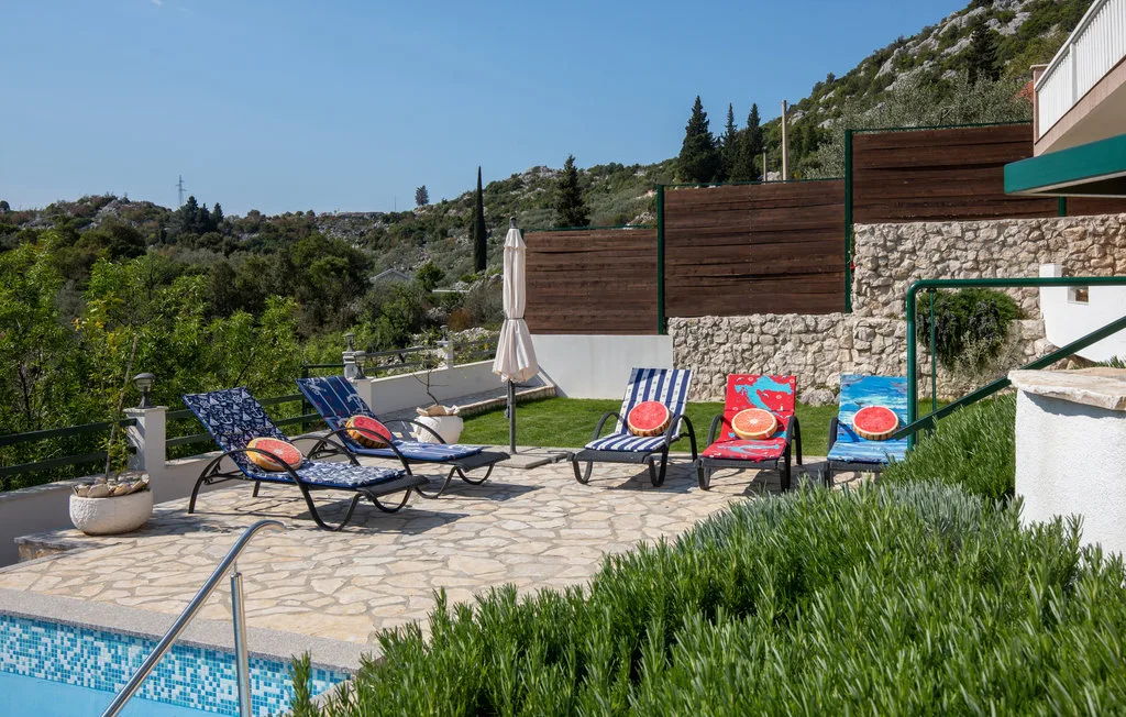 Un patio soleado junto a la piscina tiene asientos reclinables, cojines coloridos y vistas a las colinas.