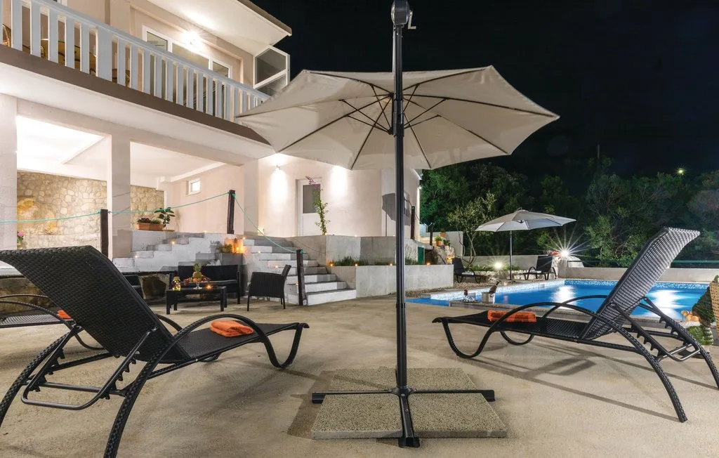 Patio nocturno con piscina, asientos reclinables y una sombrilla grande.