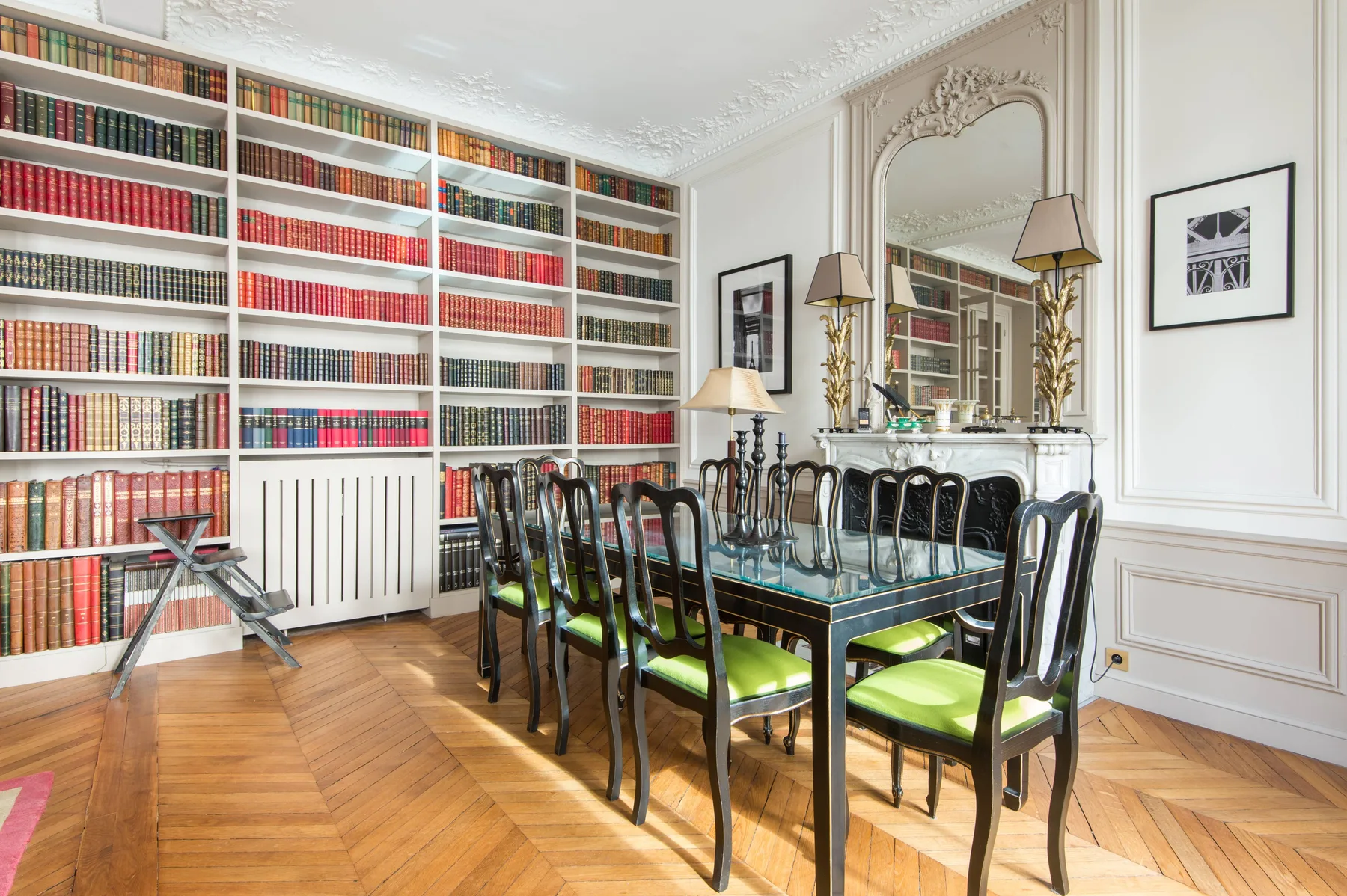 A dining table with green seating stands in a bright room lined with bookshelves.