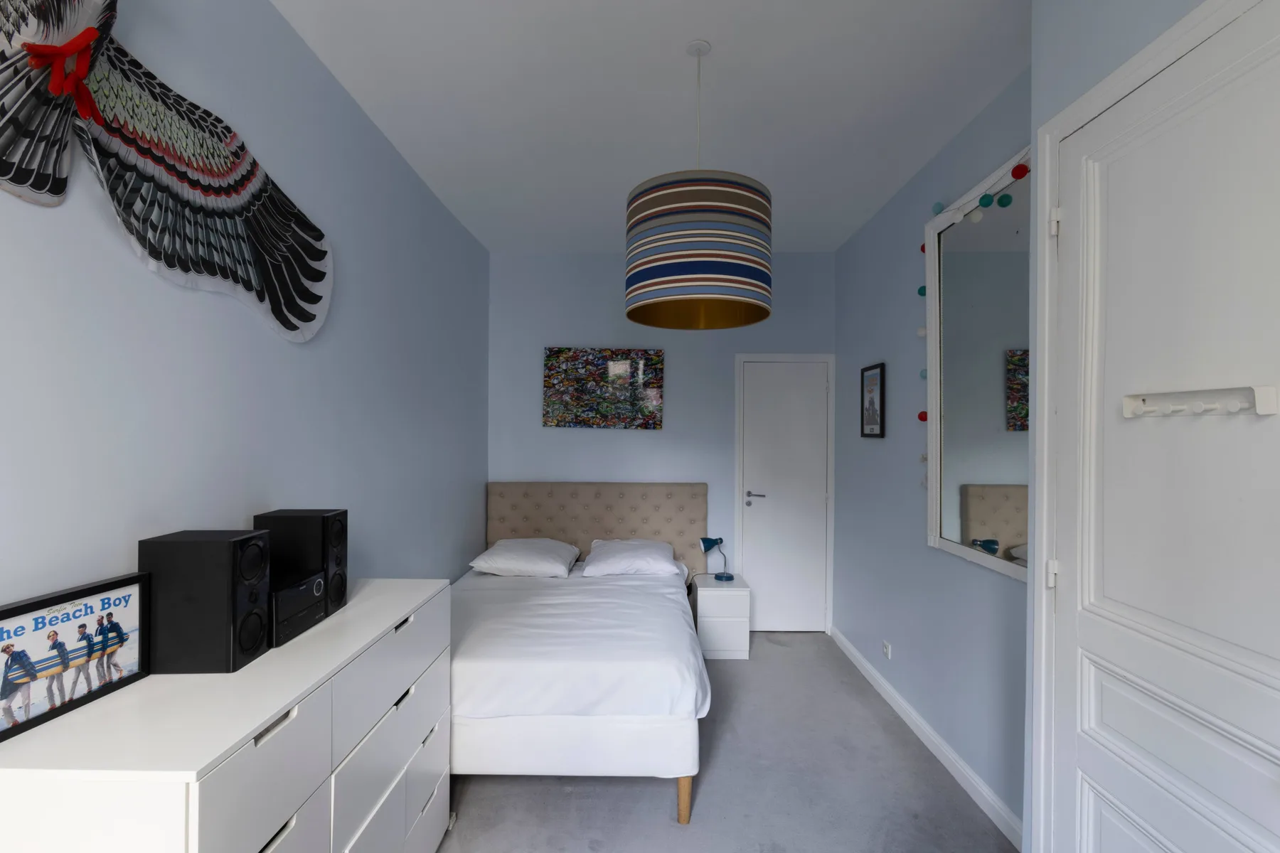 A bedroom with a neatly made bed, a white dresser, and pale blue walls.