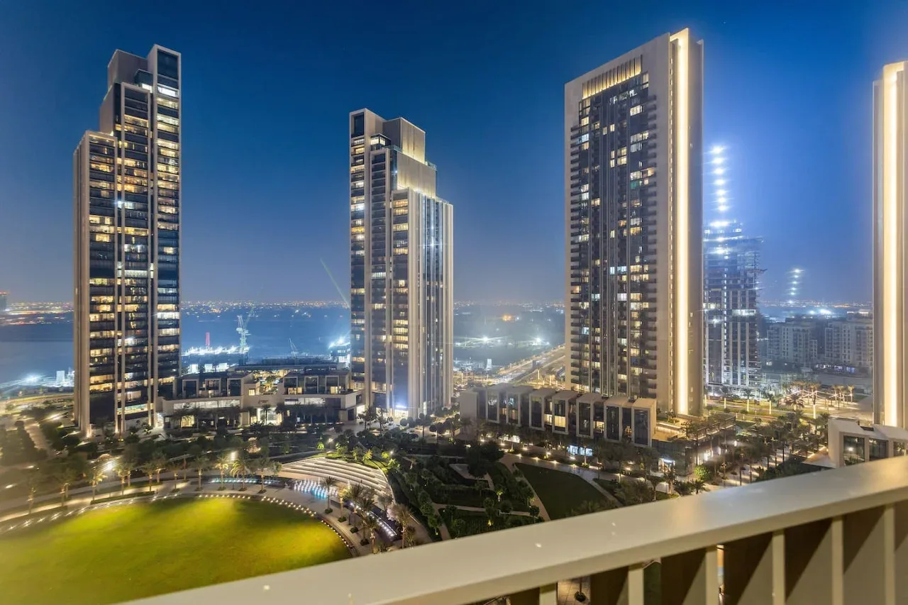 A nighttime cityscape shows illuminated high-rise buildings and landscaped grounds.