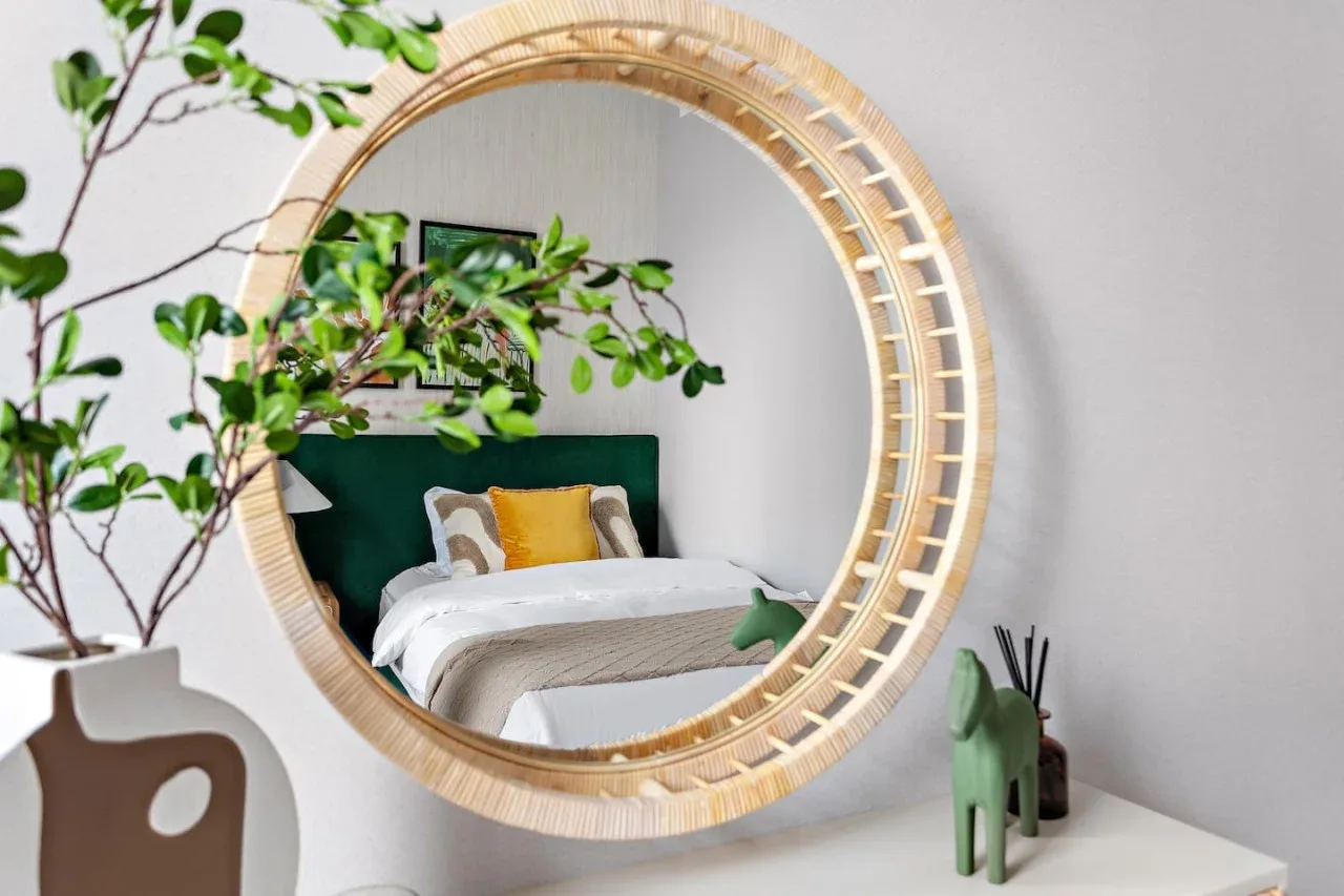 A bedroom is reflected in a large round mirror, with a bed and leafy branches.