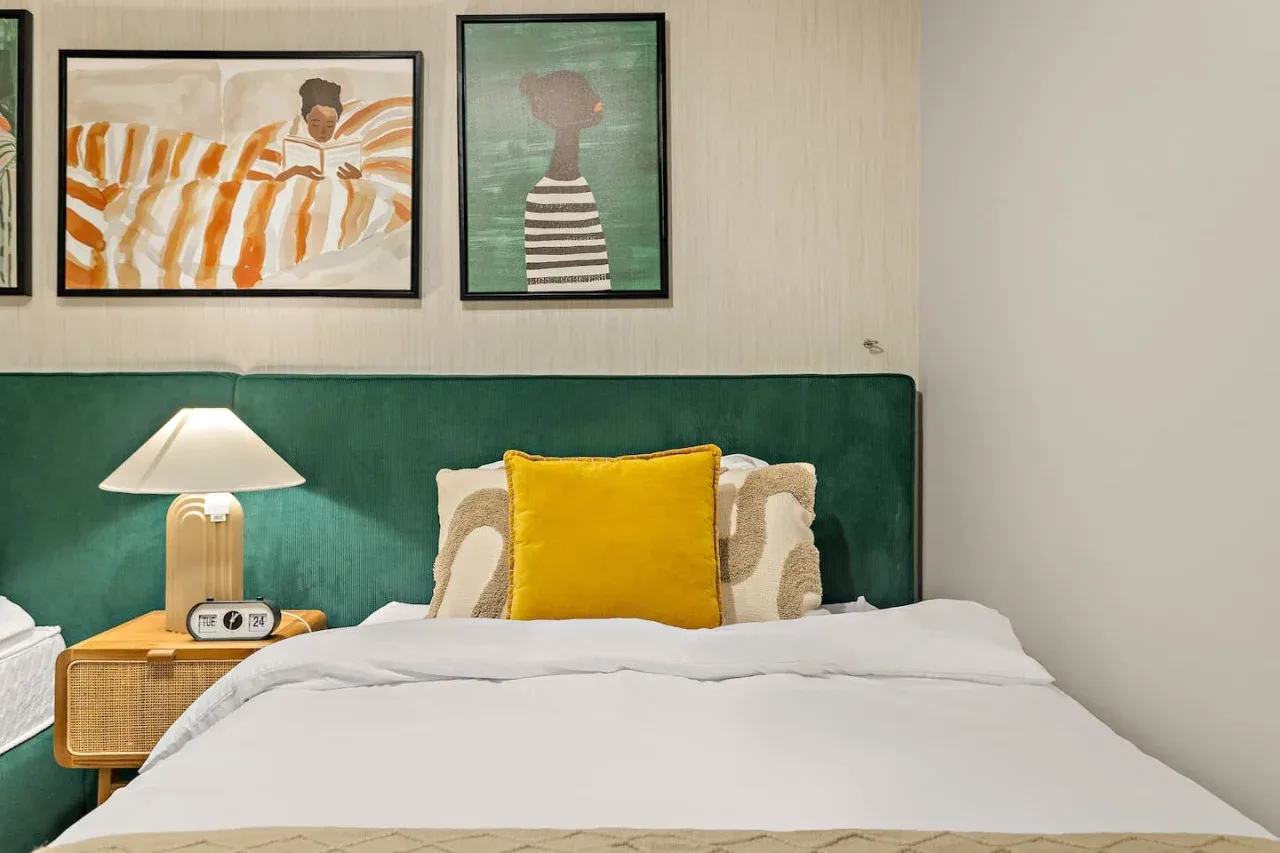 A bedroom with white bedding, a yellow accent pillow, and framed artwork above the bed.