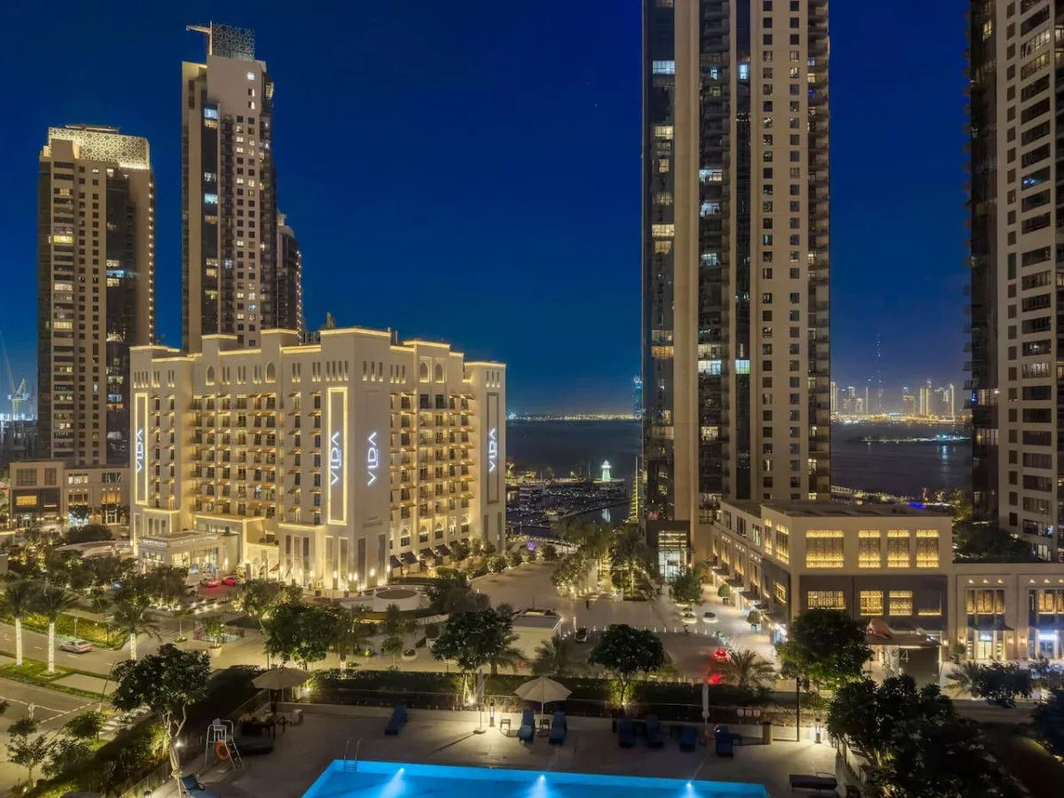 A nighttime cityscape shows illuminated high-rise buildings around a landscaped courtyard and pool.