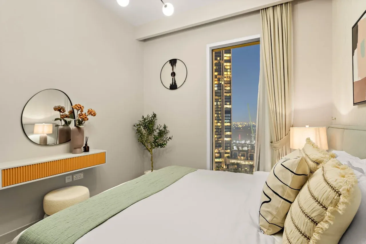 A bedroom with a bed, bedside lamp, and a large window overlooking city lights.