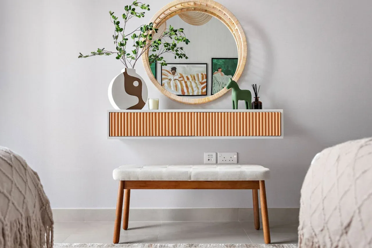 A bedroom seating area features a wooden bench beneath a round mirror and floating shelf.