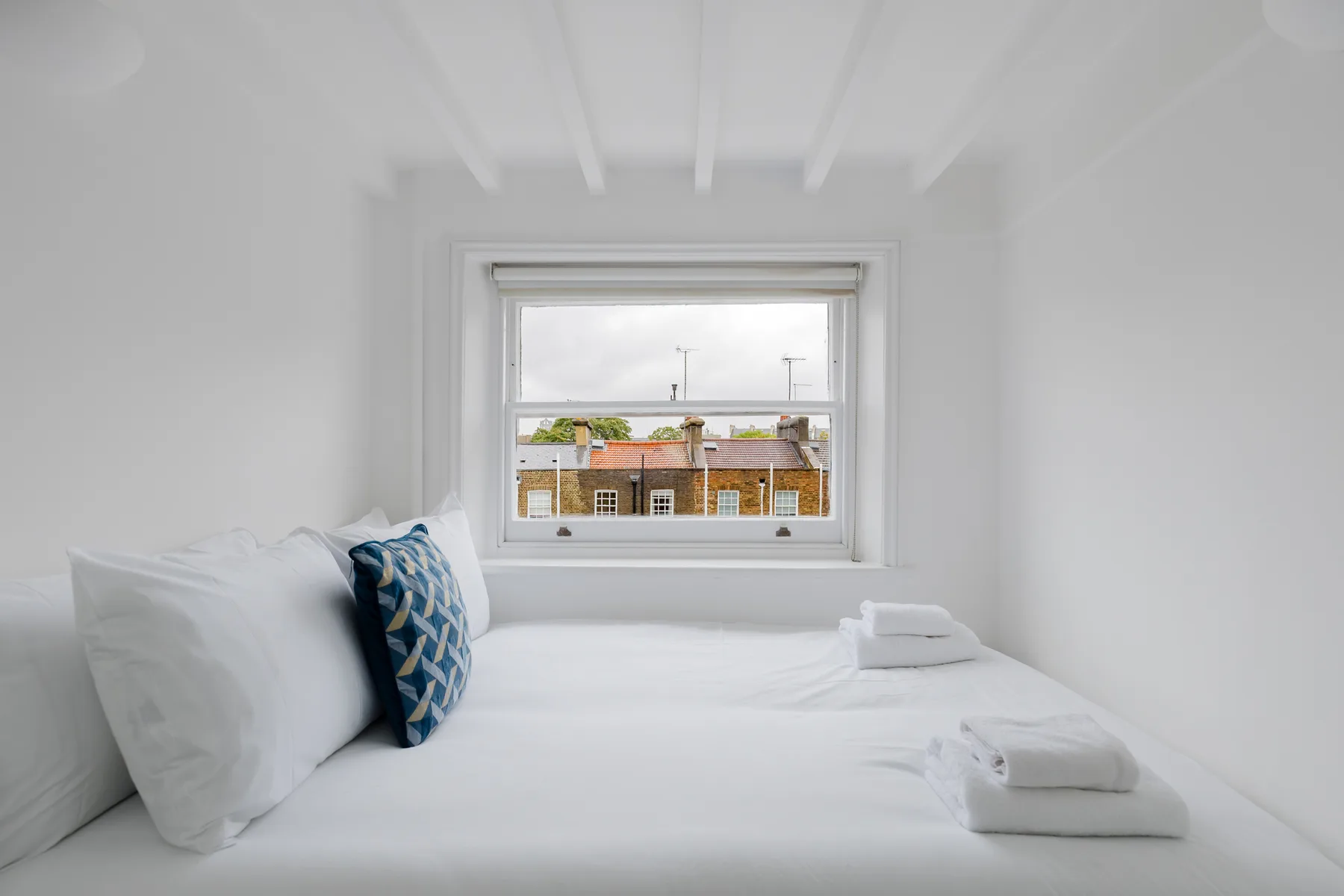 Un dormitorio luminoso con ropa de cama blanca y una ventana con vista a edificios cercanos.