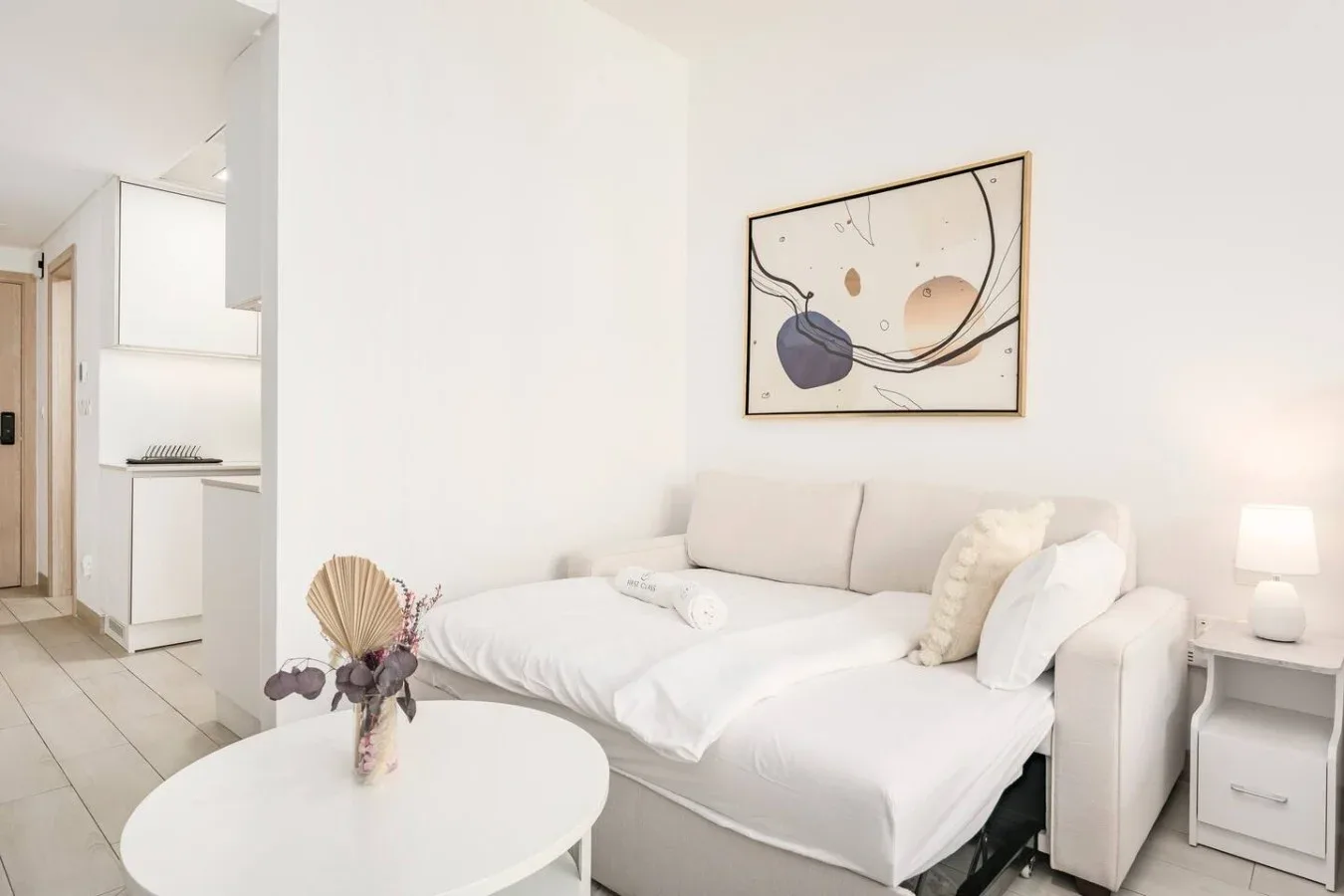 A bright bedroom with white bedding, a seating area, and abstract wall art.