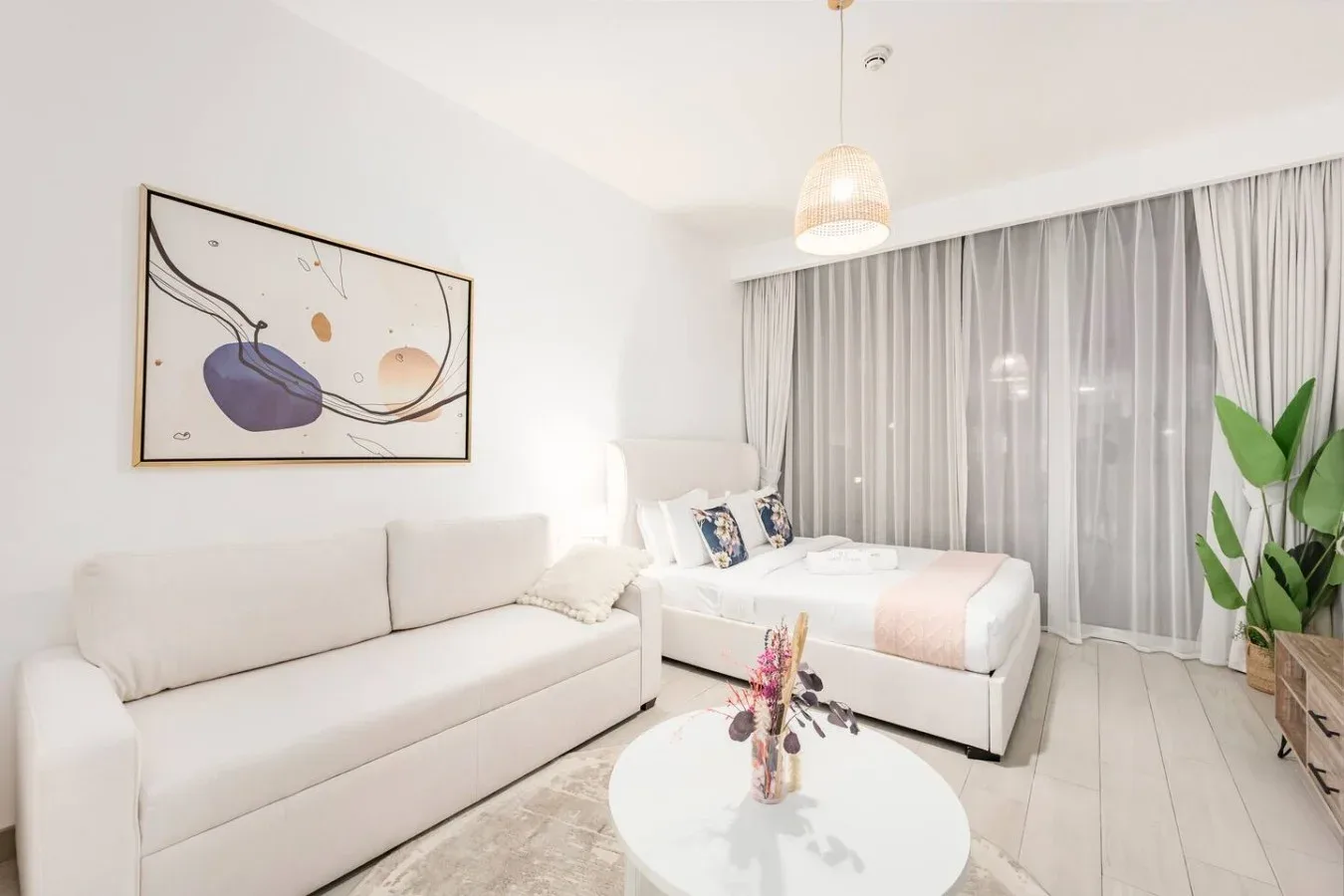 A bright studio room with a sofa, bed, and large curtained window.