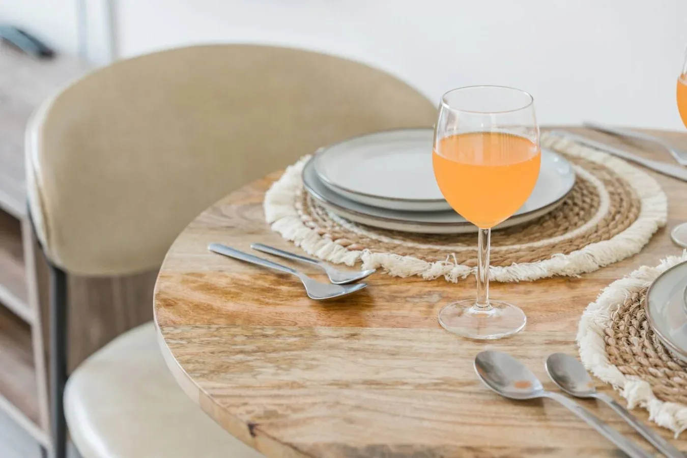 A dining table is set with plates, woven placemats, cutlery, and a glass of orange juice.