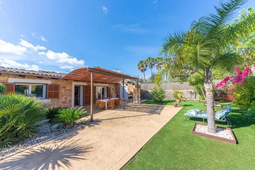 A sunny backyard patio beside a brick house, with a lawn and palm trees.