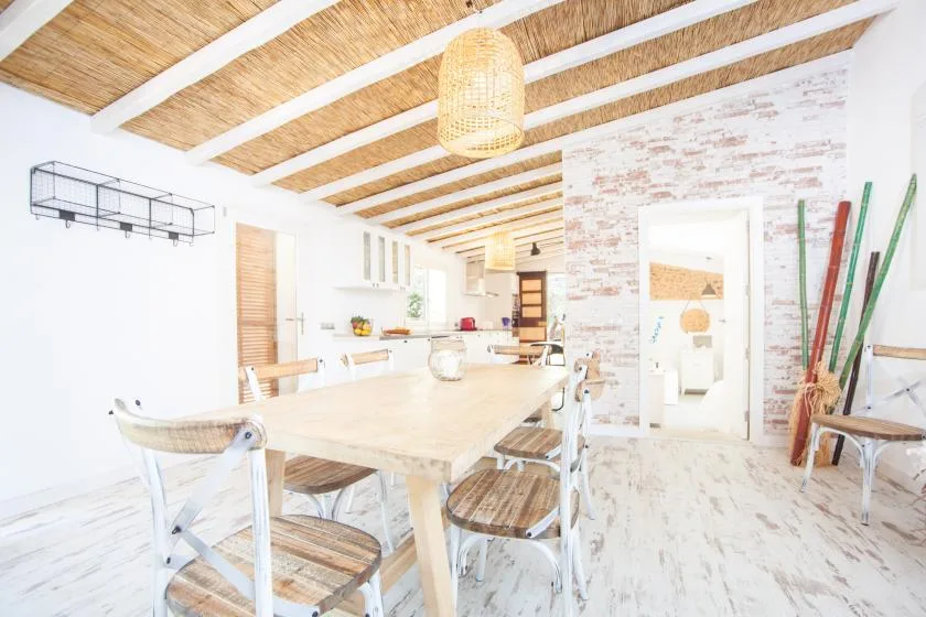 A bright dining area with a long table and chairs beneath exposed ceiling beams.