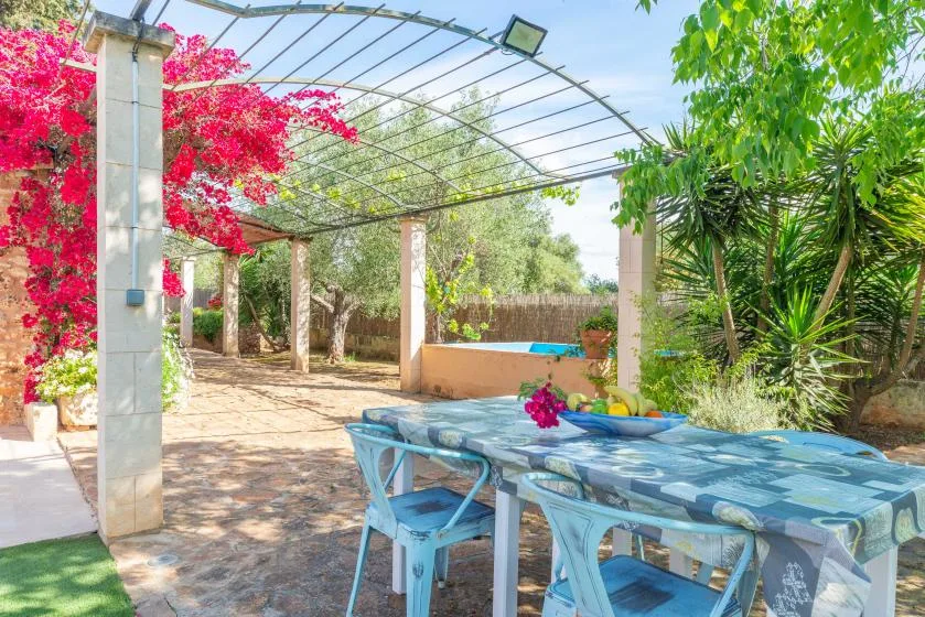 A shaded outdoor dining area sits beneath a pergola beside lush greenery.