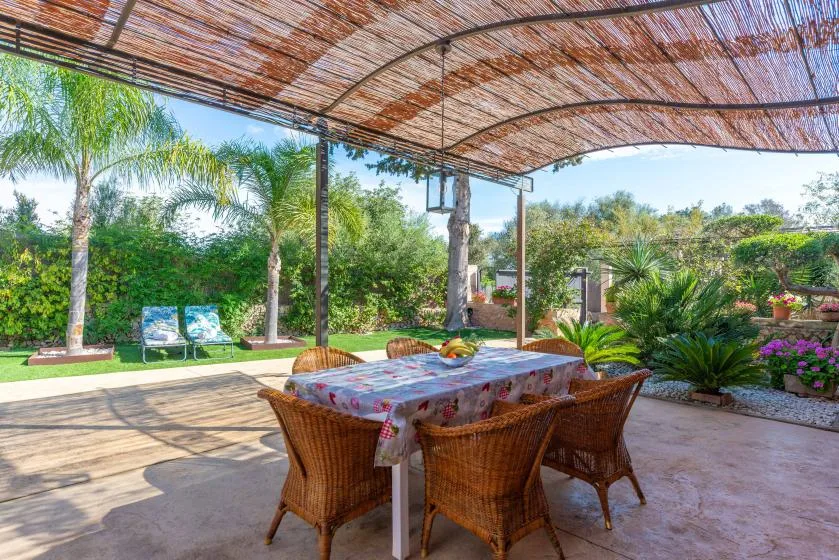 An outdoor dining area sits beneath a shaded canopy, surrounded by a garden.