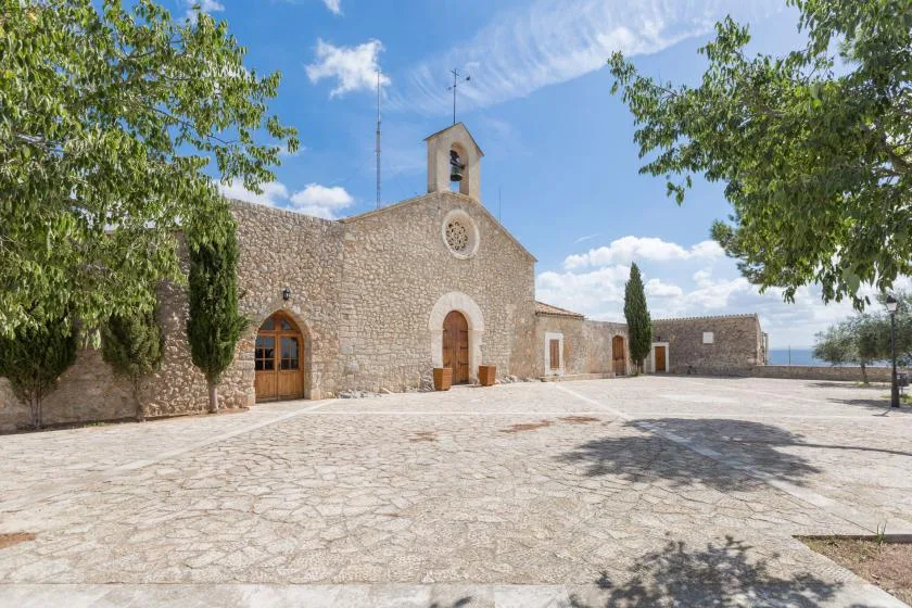 A stone chapel faces a wide paved courtyard beneath a blue sky.