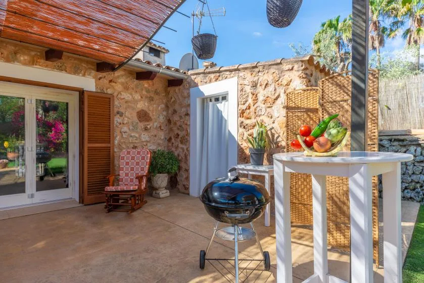 A sunlit stone patio with a barbecue grill and a white table holding fruit.