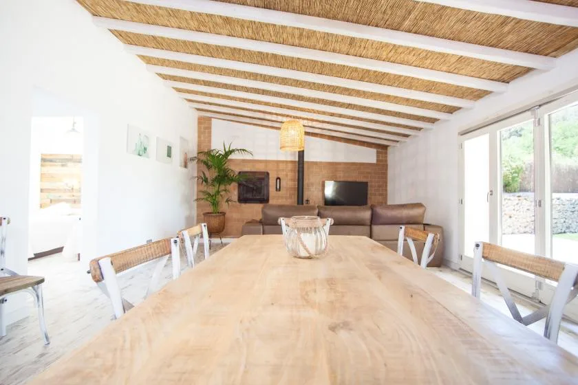 A bright dining and living area features a long wooden table and seating beneath exposed ceiling beams.