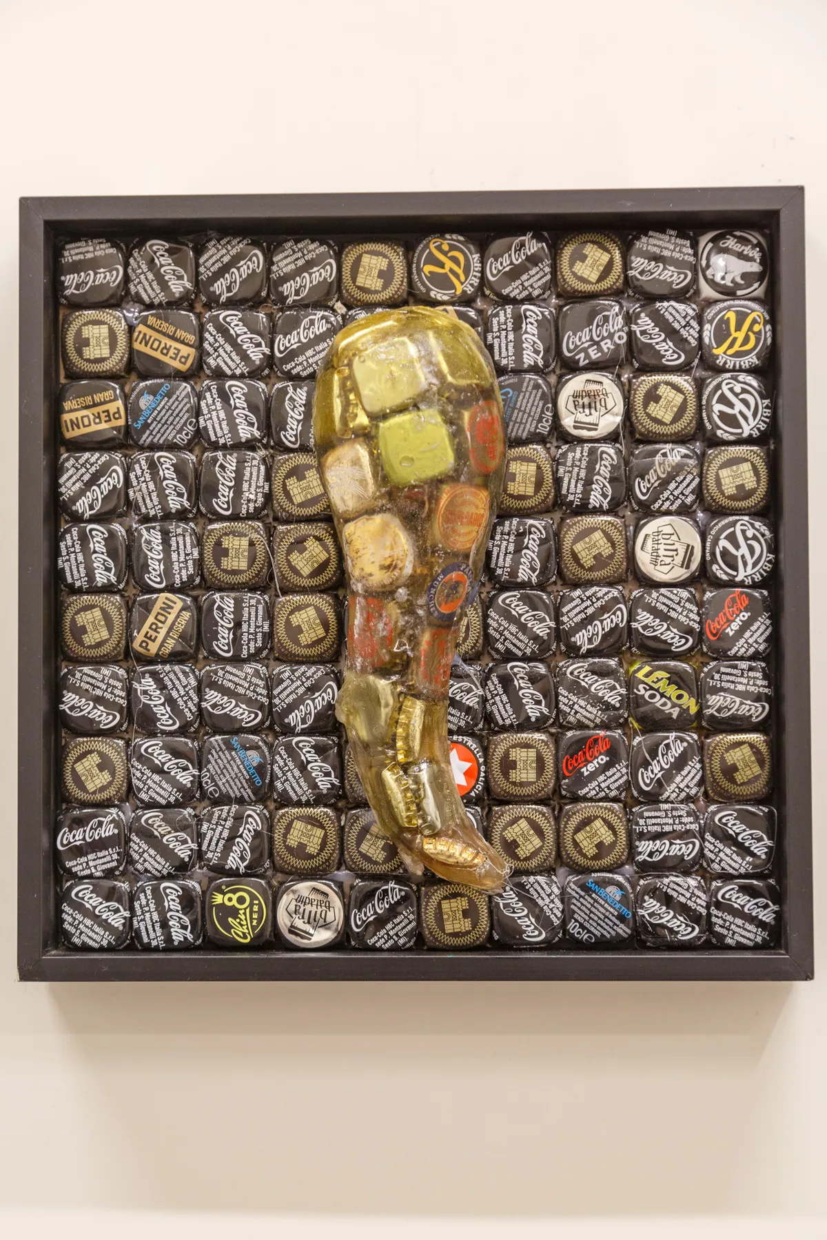 Una obra enmarcada en la pared muestra una figura dorada sobre un fondo de tapas de botellas.